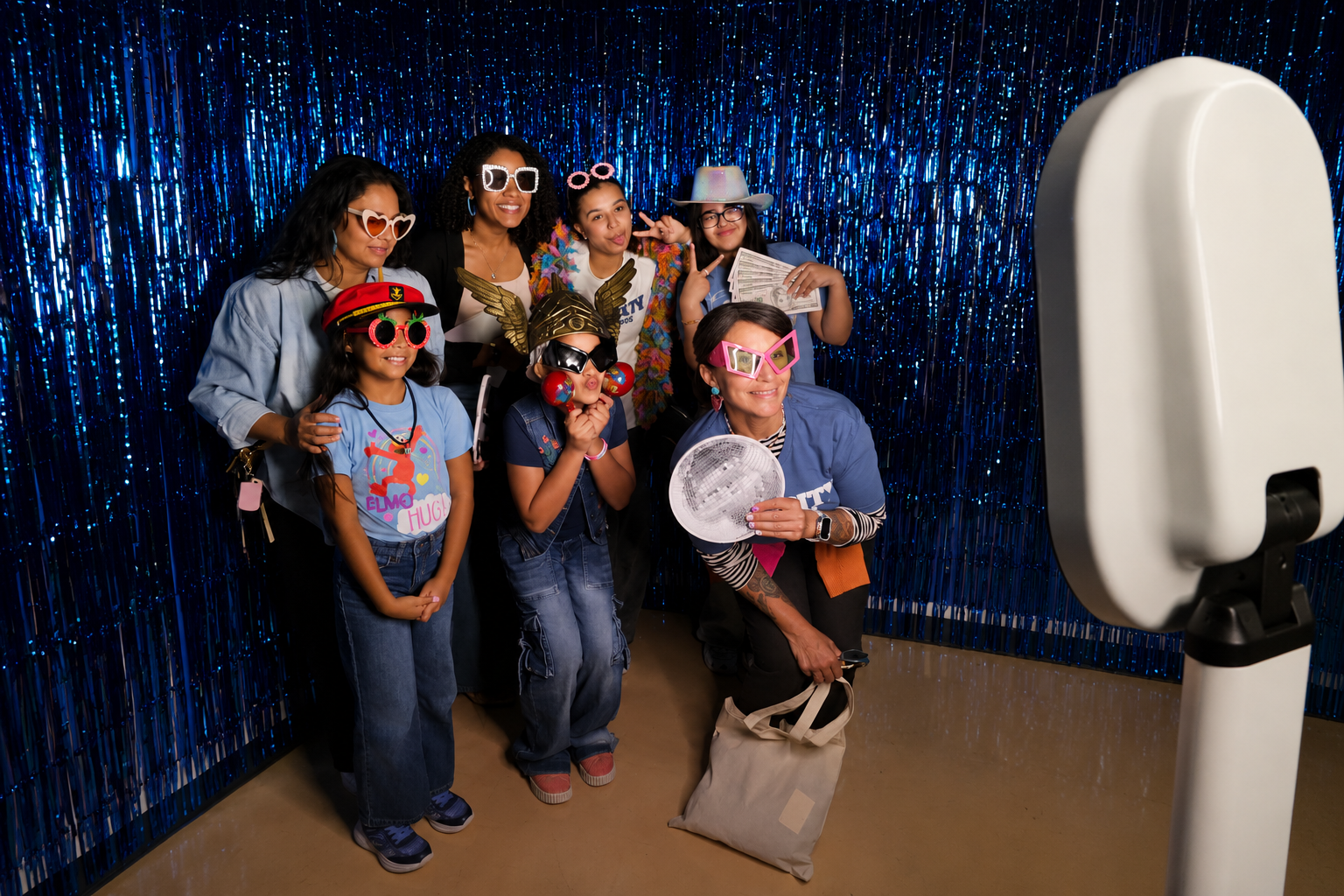 Group of guests posing with colorful props and novelty glasses in front of a shimmering blue tinsel backdrop at a photo booth event setup.