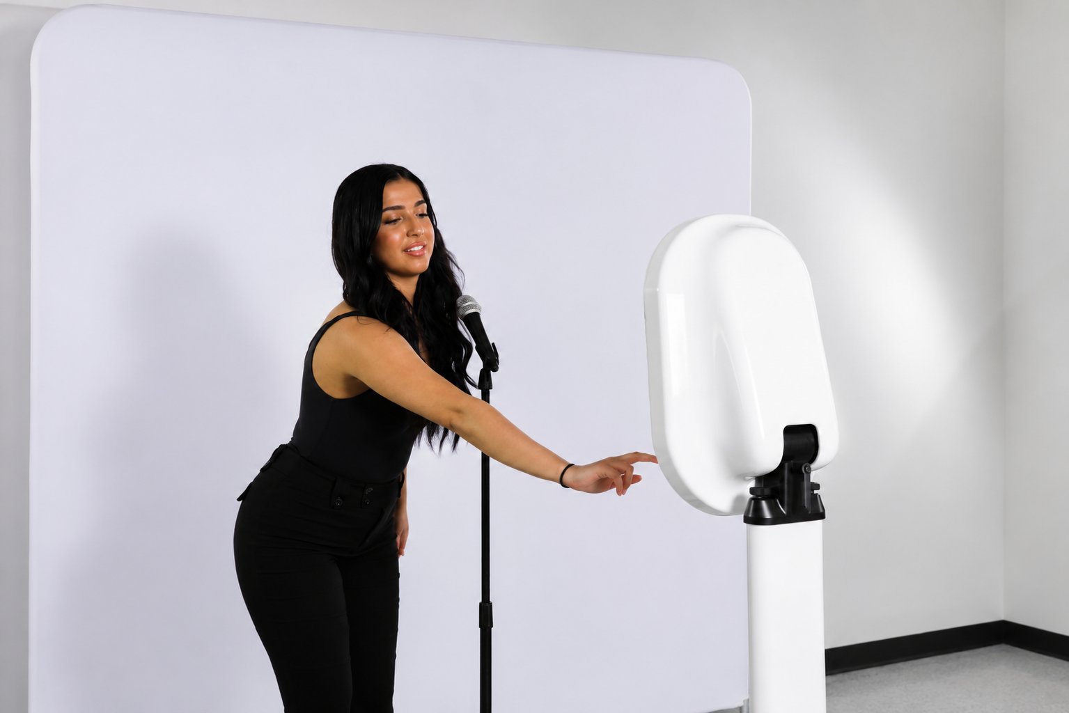 
Woman interacting with a modern white photo booth beside a microphone stand in a minimalist studio setup with a clean white backdrop.
